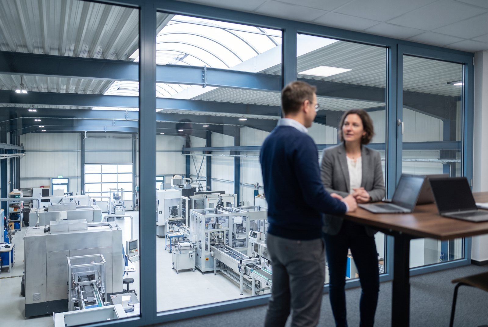 Zwei Personen im Gespräch an einem Stehtisch, dahinter eine Produktionshalle